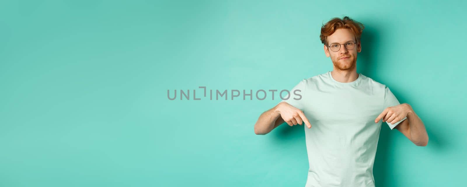 Skeptical and bothered man with ginger hair and beard, wearing glasses and t-shirt, smirk and point fingers down, showing promo with judgy face, turquoise background.