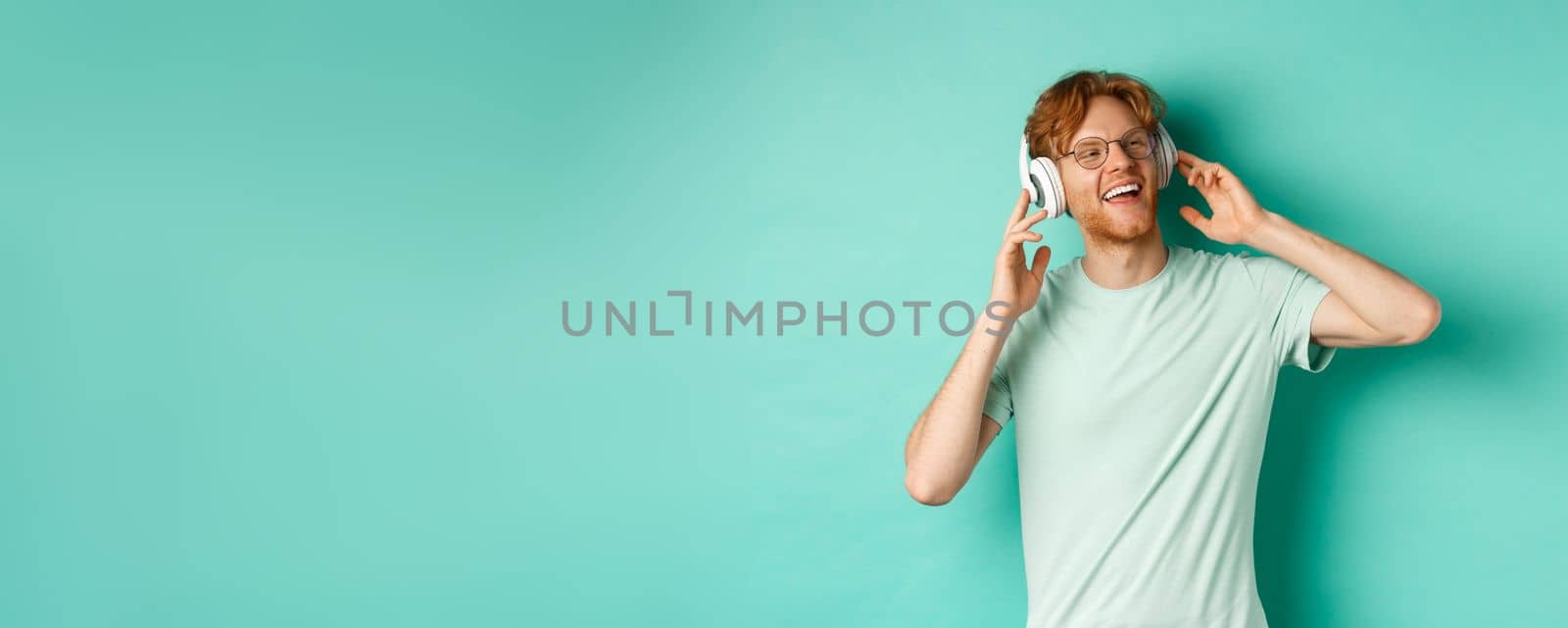 Lifestyle concept. Happy young man with ginger hair dancing and having fun, listening music on wireless headphones and smiling pleased, turquoise background by Benzoix