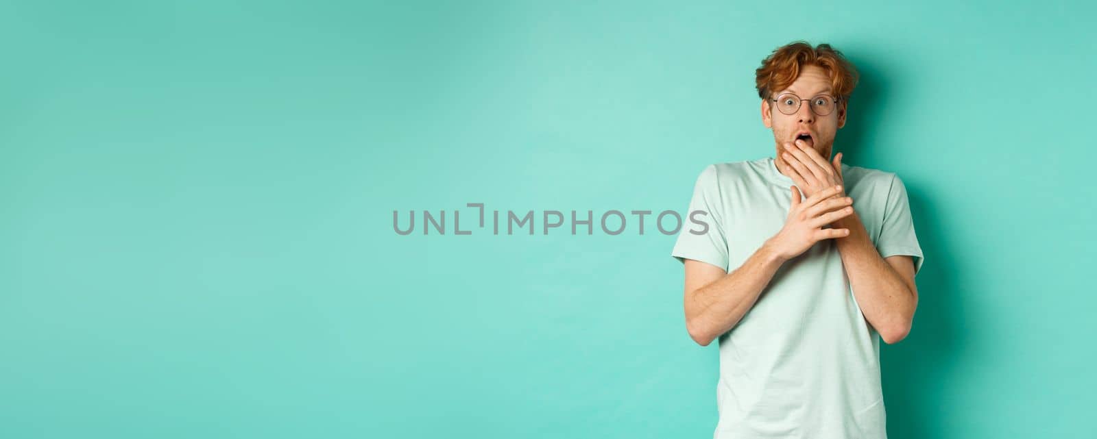 Shocked redhead man in glasses gasping startled, covering mouth and staring at camera scared, standing over turquoise background.
