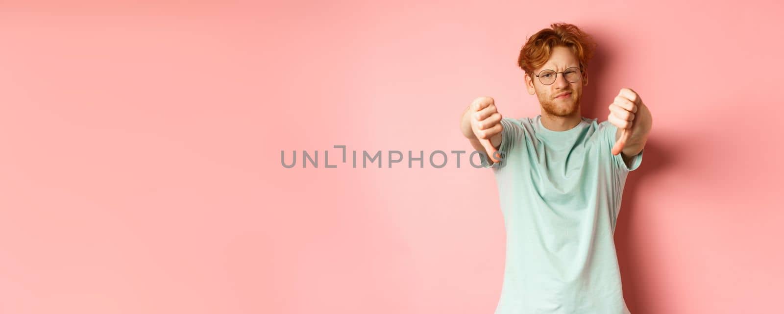 Disappointed young man in glasses, with messy red haircut, showing thumbs down and grimacing unsatisfied, express dislike, standing over pink background.