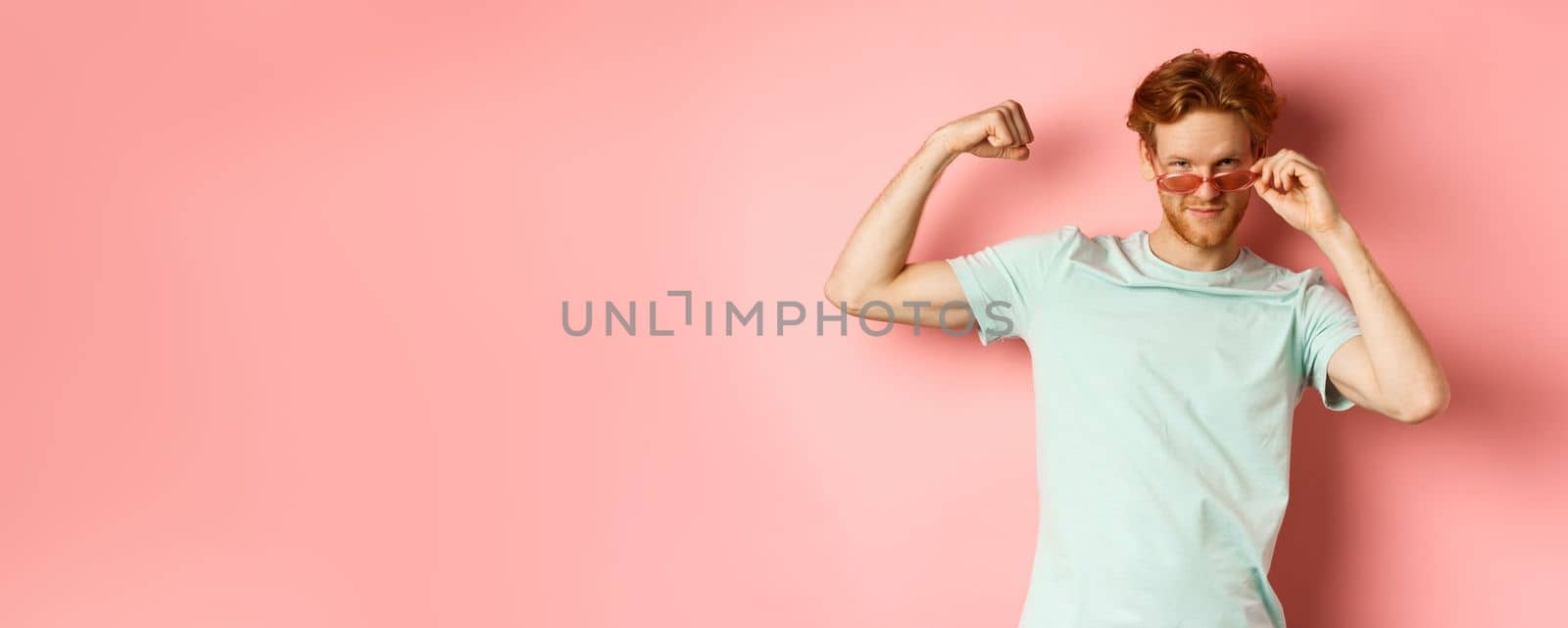 Confident young man with red hair, wearing summer sunglasses and t-shirt, showing strong and fit body muscles, flex biceps and staring cool at camera, pink background by Benzoix