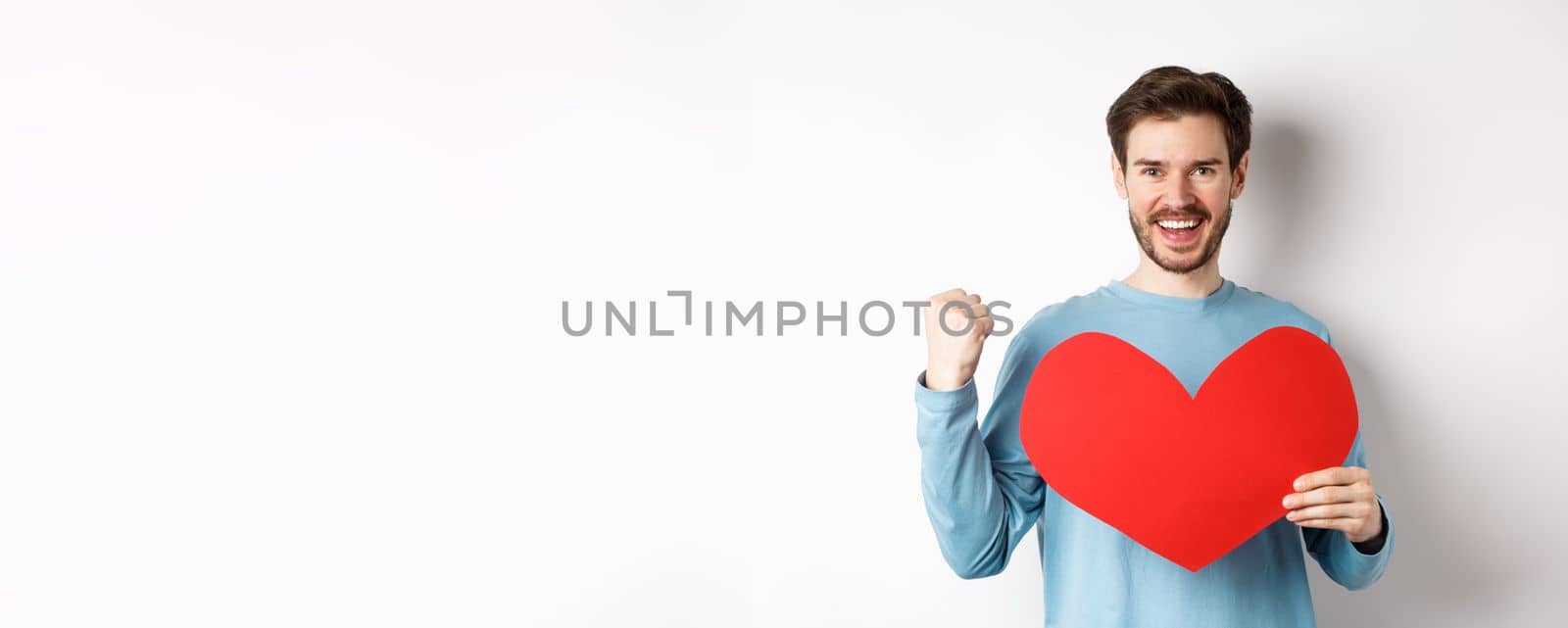 Cheerful man determined to find love on Valentines day, making fist pump success gesture and holding big red romantic heart, standing over white background.