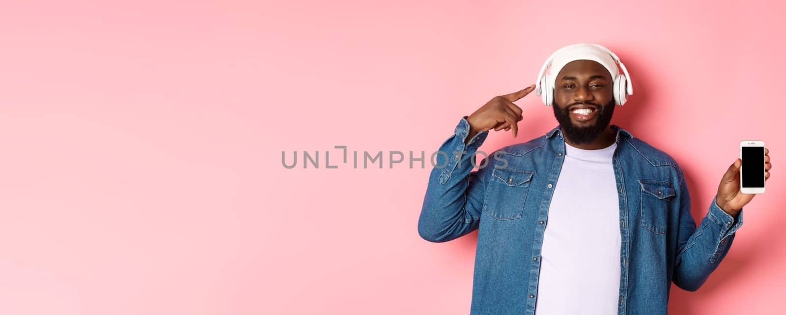 Happy black man listening music, pointing finger at headphones and smiling, showing mobile phone screen app or playlist, standing over pink background by Benzoix