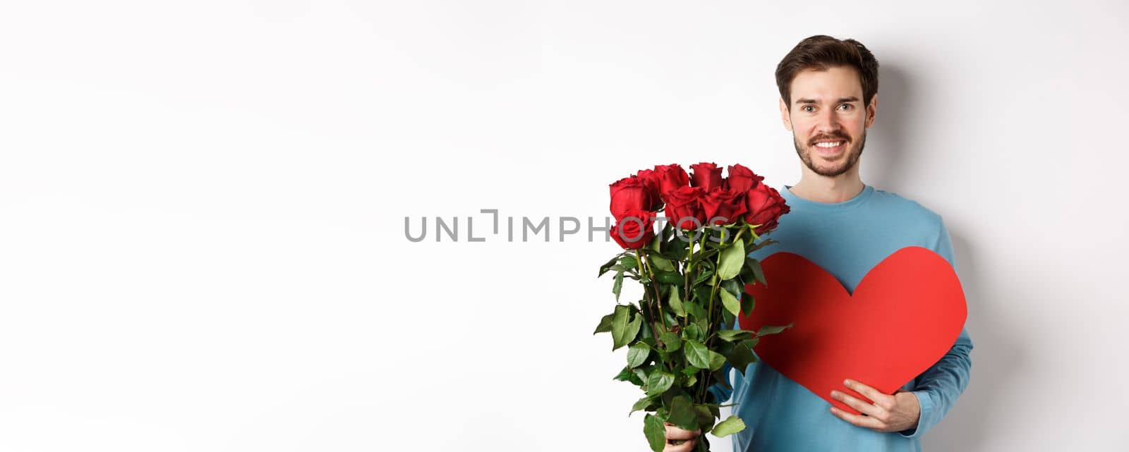 Romantic people. Handsome smiling man holding bouquet of roses and big red heart, going on Valentines day date with girlfriend, standing over white background.
