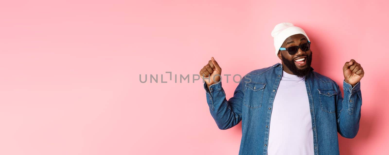 Happy and carefree Black man celebrating, dancing and having fun, wearing beanie and sunglasses, standing over pink background.