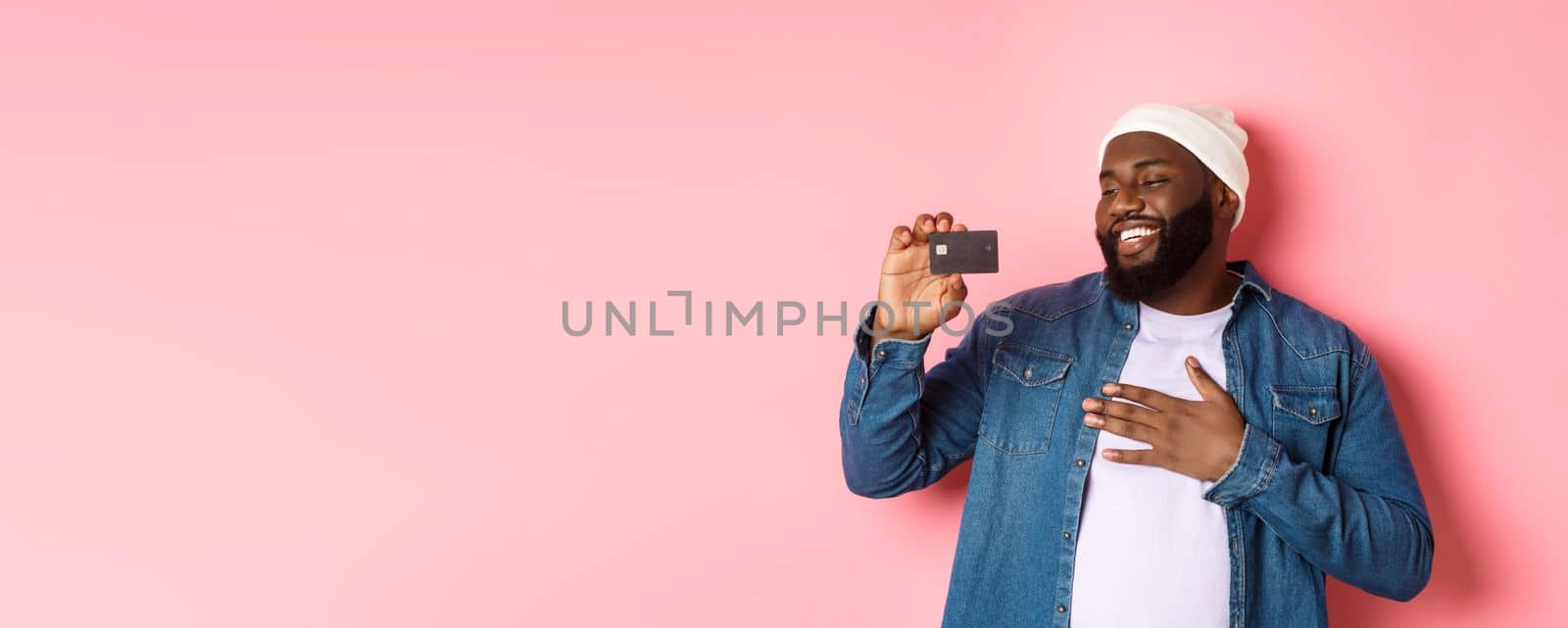 Shopping concept. Image of happy african-american man looking satisfied at credit card, recommending bank, standing over pink background.