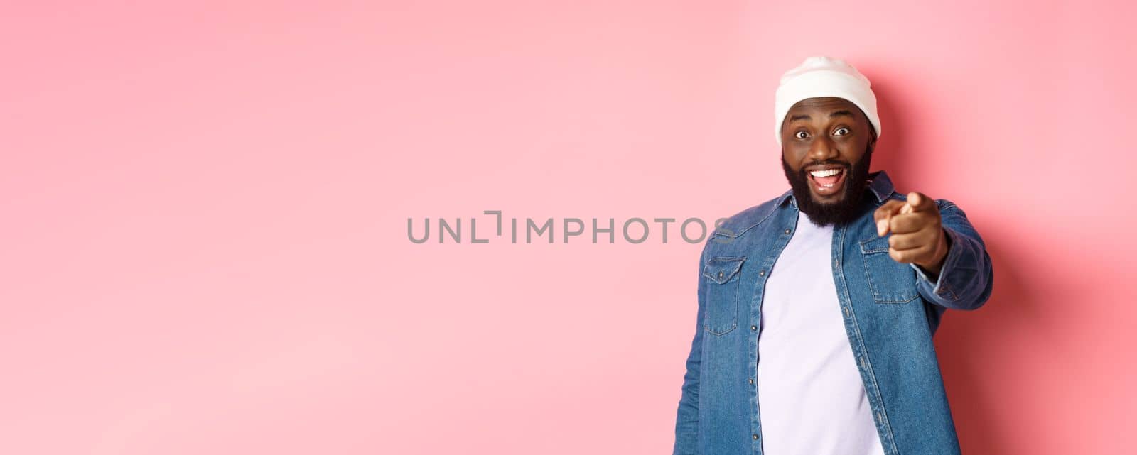 Happy african-american hipster man pointing finger at camera, need you, smiling excited, standing over pink background.