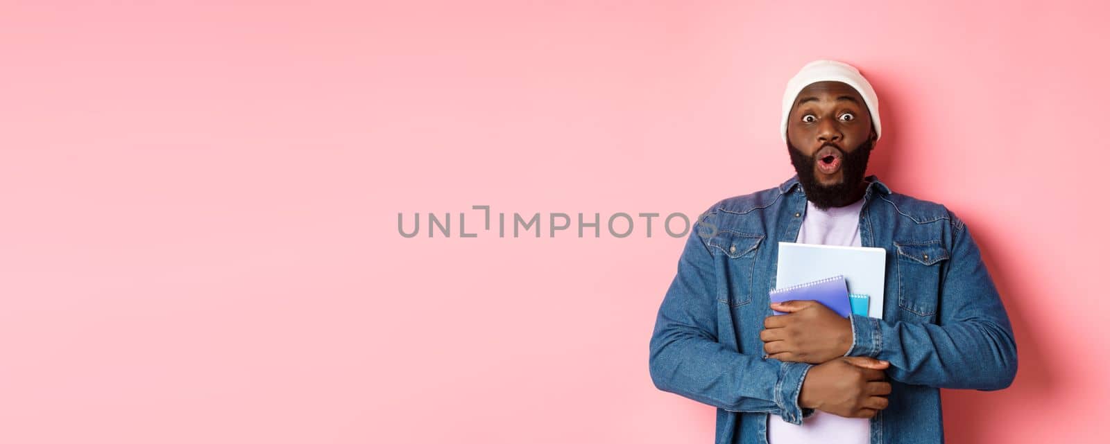 Education. Excited african-american adult student carry notebooks, staring at camera amaze, standing over pink background.