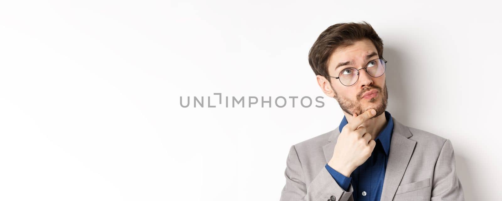 Thoughtful businessman with beard and glasses making decision, looking up and thinking, standing in suit with pensive face, white background.