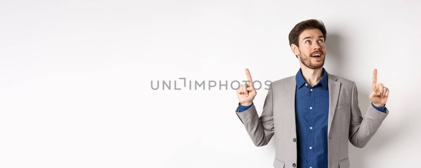 Excited handsome man with beard, wearing suit and looking amused up, pointing at top with admiration, standing against white background.