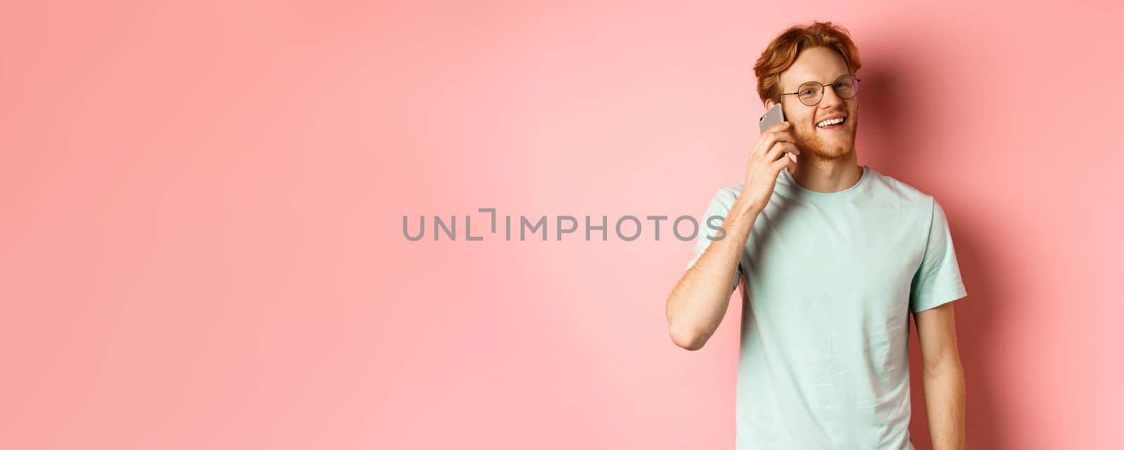 Handsomy hipster guy with red hair and beard talking on mobile phone, calling someone and looking happy, standing over pink background by Benzoix