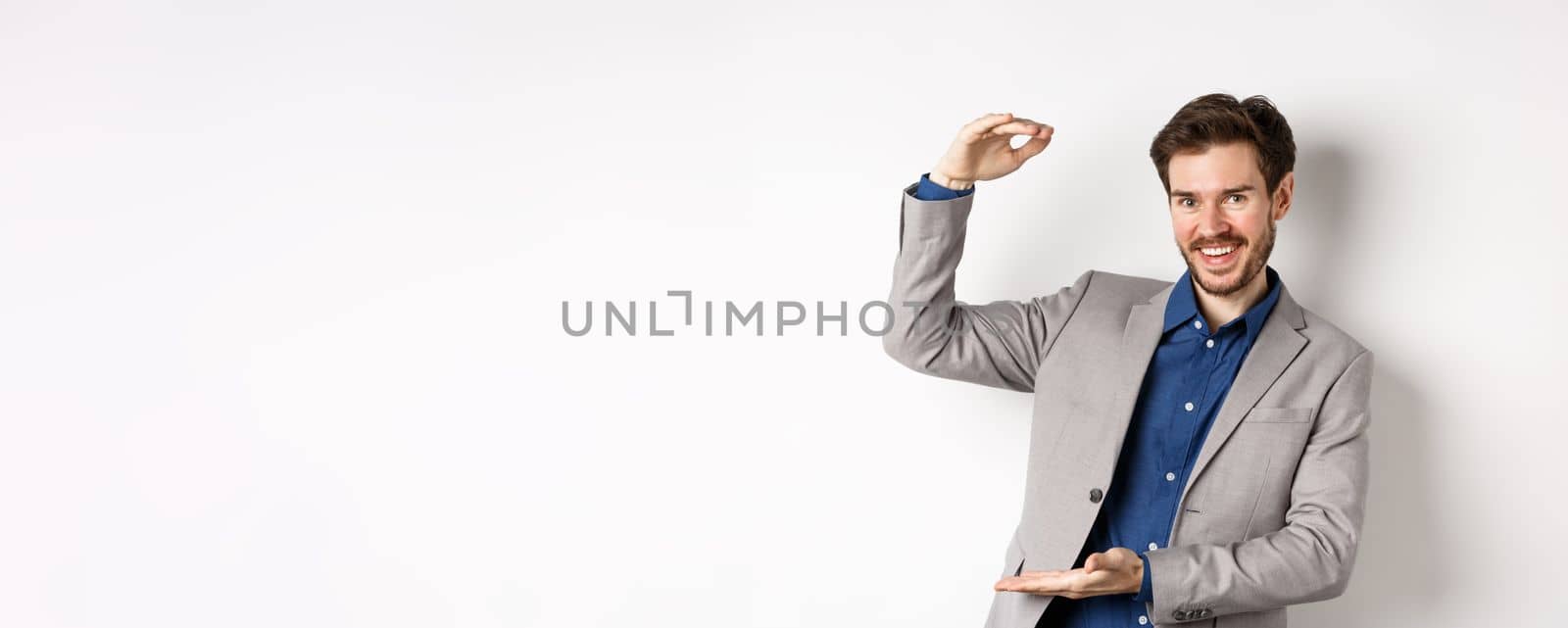 Successful businessman showing big size, shape large object and smiling pleased, standing on white background in suit.