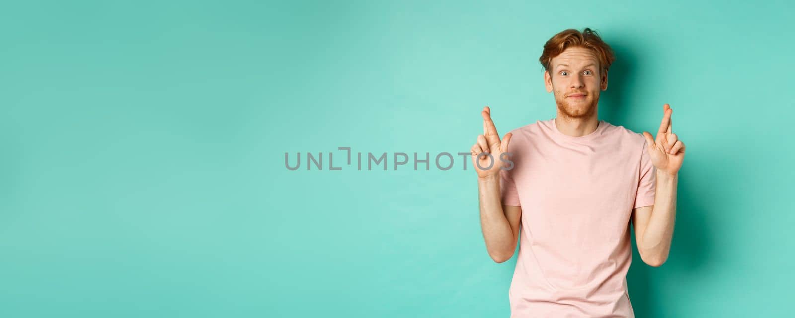 Smiling hopeful man with red hair making a wish, cross fingers for good luck and expecting something good, standing over turquoise background.