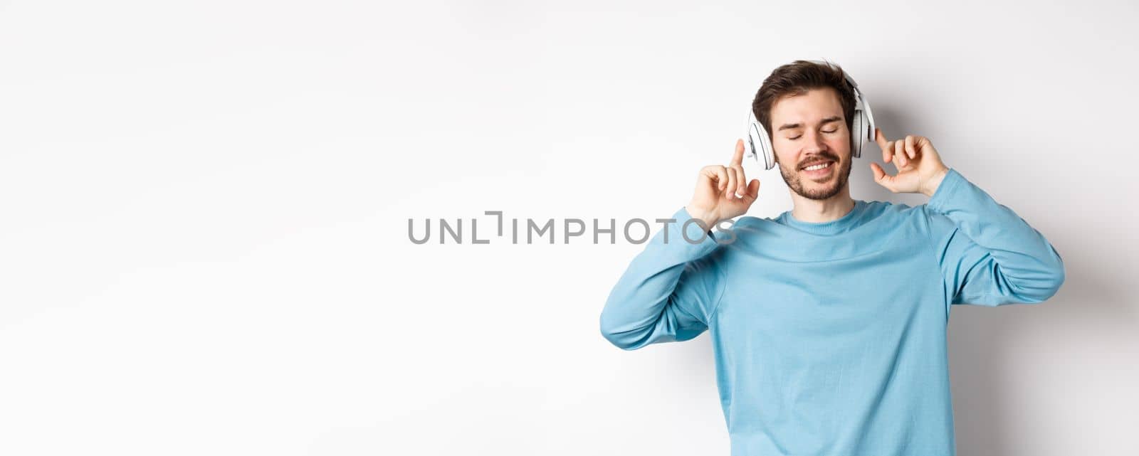 Handsome man listening music in wireless headphones and smiling, enjoying good sound, white background.