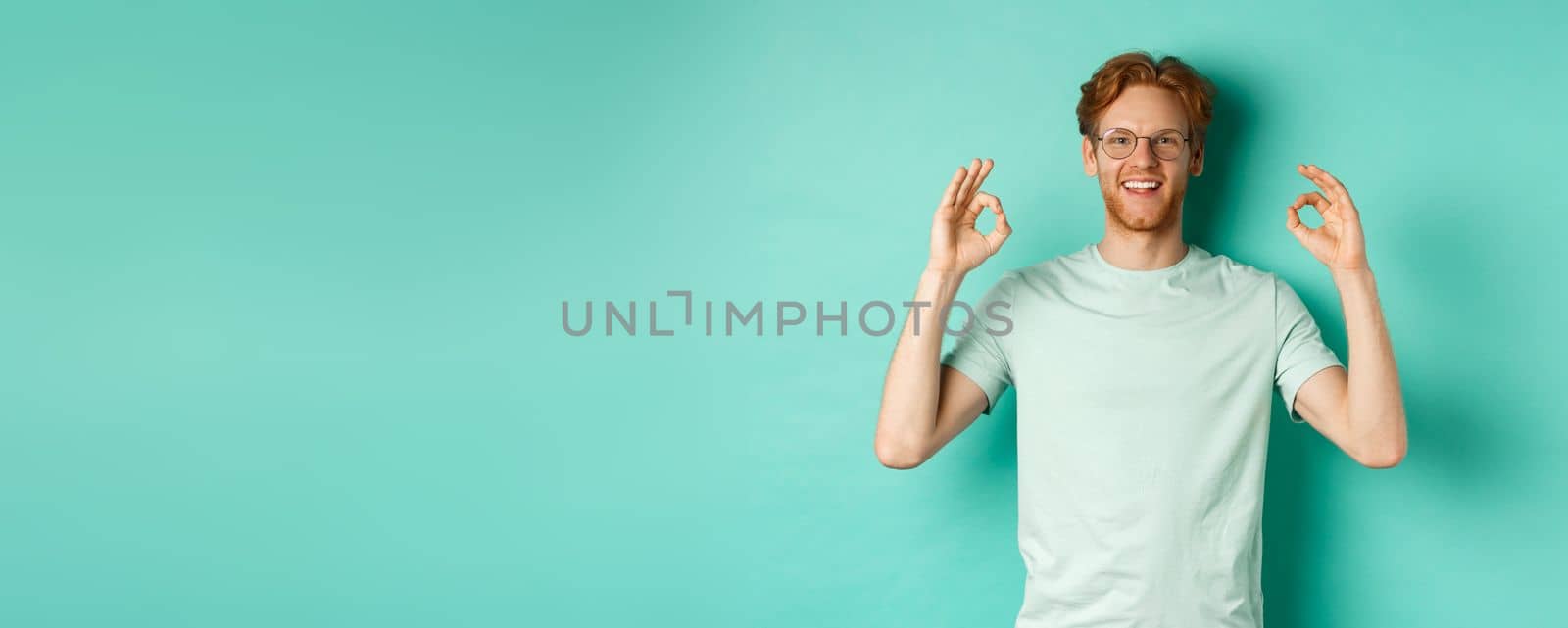 Attractive male model with red hair, wearing glasses, showing OK sign in approval and saying yes, smiling satisfied, standing over mint background.