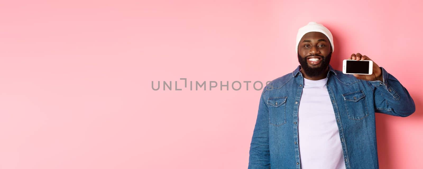 Online shopping and technology concept. Handsome Black man showing smartphone screen horizontally and smiling satisfied, recommending app, pink background.