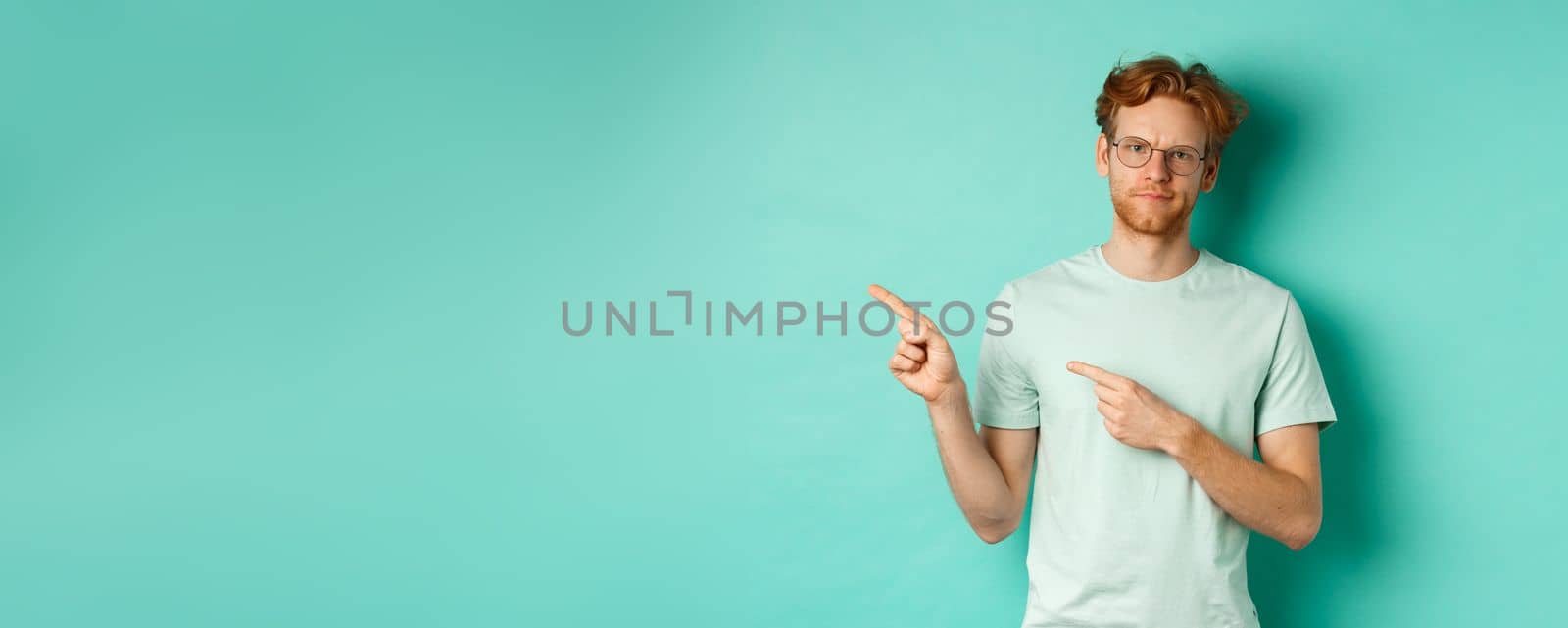 Judgemental redhead man in glasses grimacing, frowning disappointed and pointing left, showing bad promo offer, standing over turquoise background by Benzoix