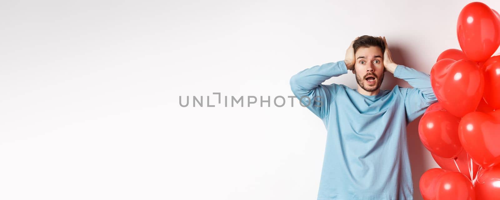 Confused boyfriend holding hands on head and panicking on Valentines holiday, alarmed with romantic presents on lovers day, standing near hearts balloon over white background by Benzoix