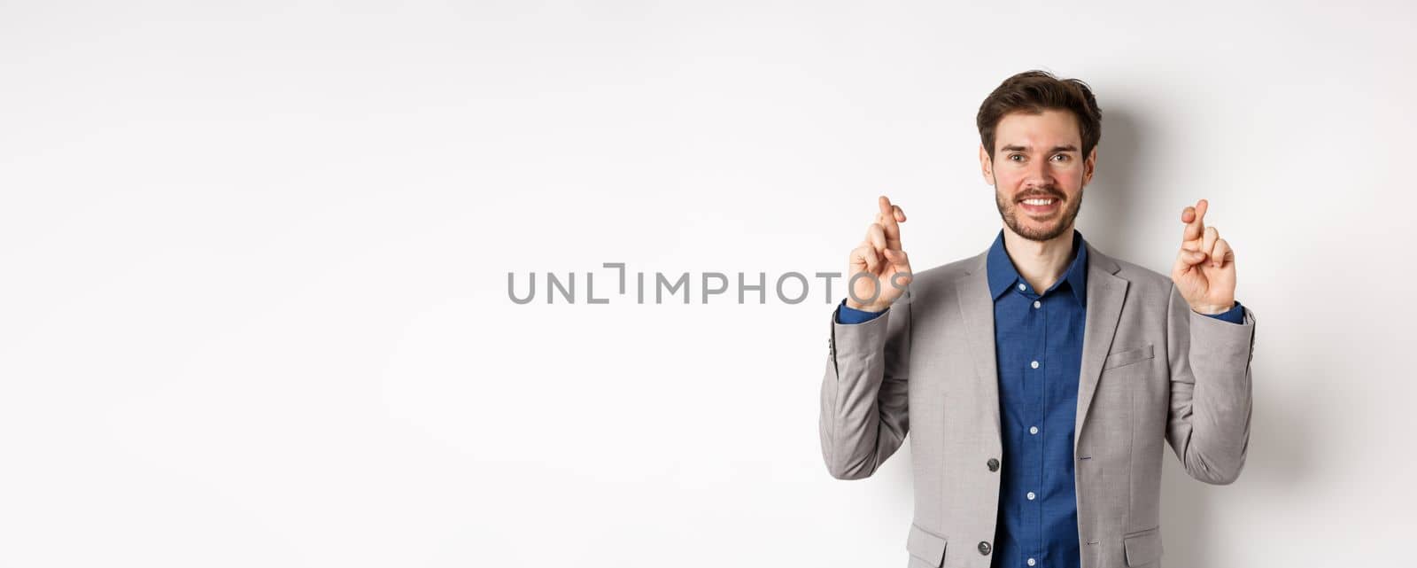 Hopeful smiling businessman looking optimistic, cross fingers good luck, praying to win, waiting for results with hope, standing on white background.