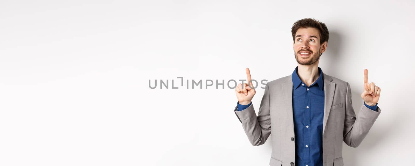 Dreamy smiling male manager in suit pointing and looking up, happy face, checking out advertisement, standing against white background.