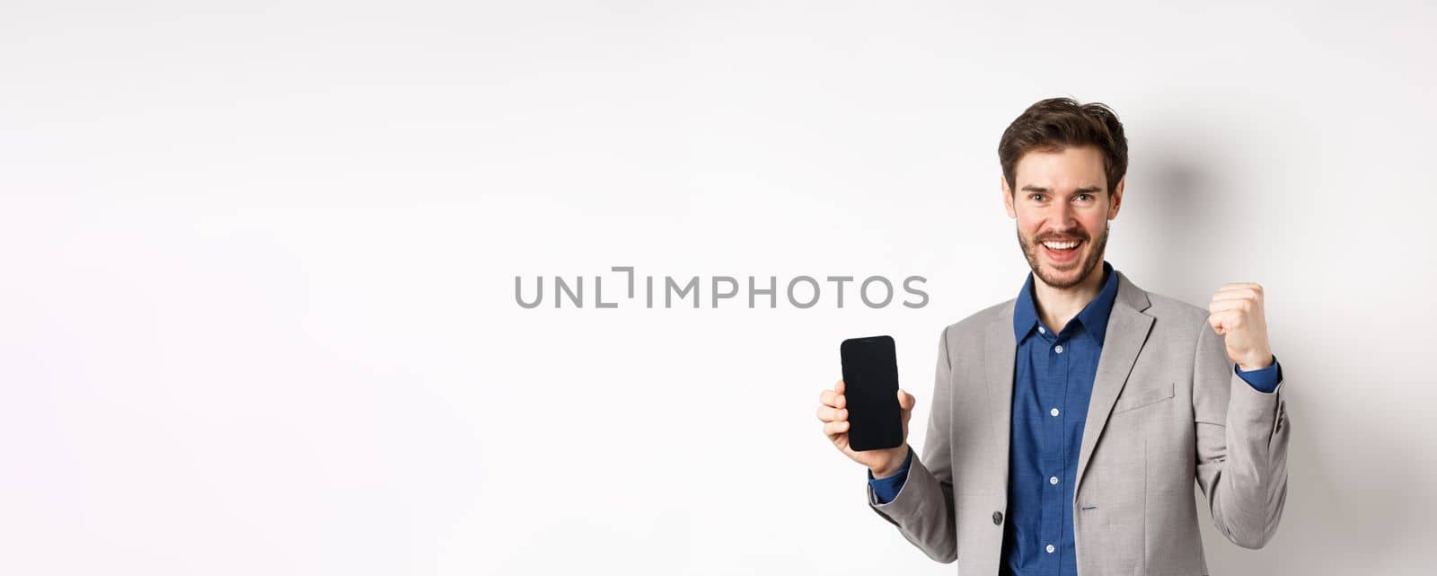 E-commerce and online shopping concept. Man making money in internet, showing smartphone screen and winner gesture, smiling satisfied, standing on white background by Benzoix