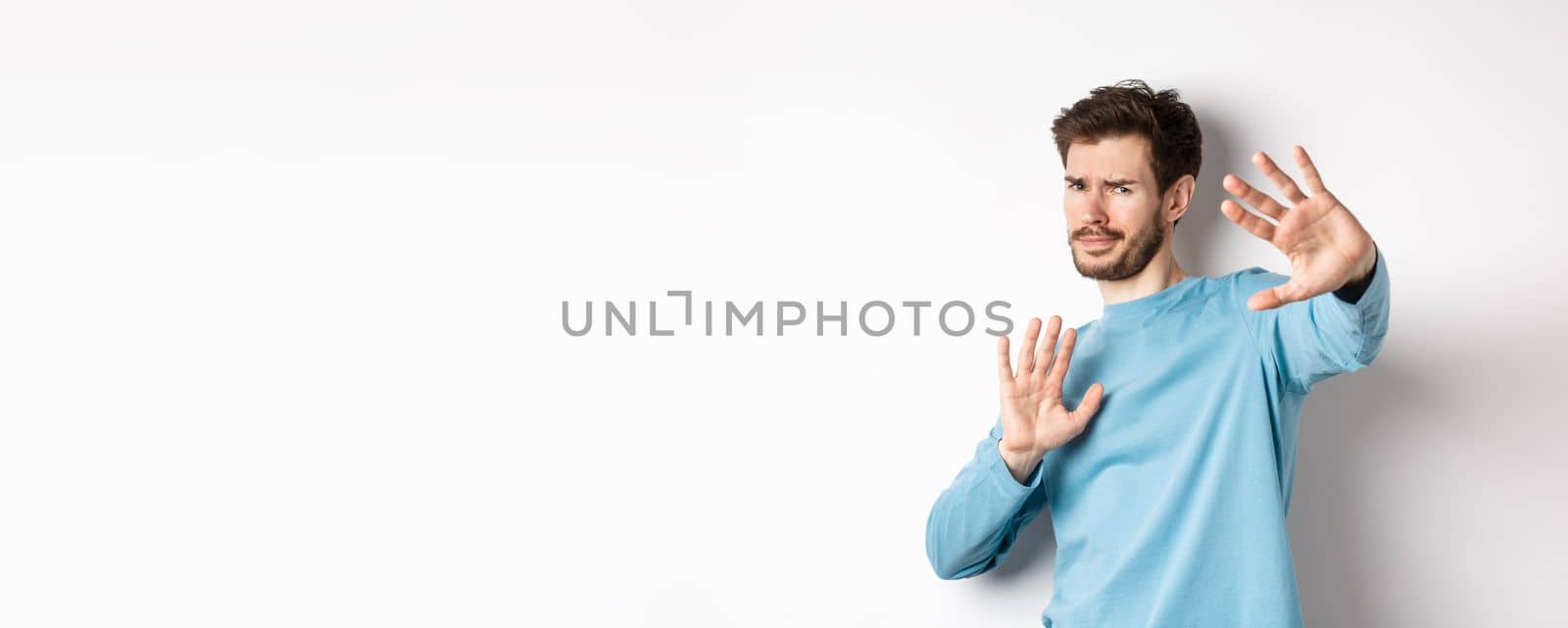 Stay away from me. Reluctant young man step back with hands stretch out in defensive gesture, protecting himself, standing over white background.