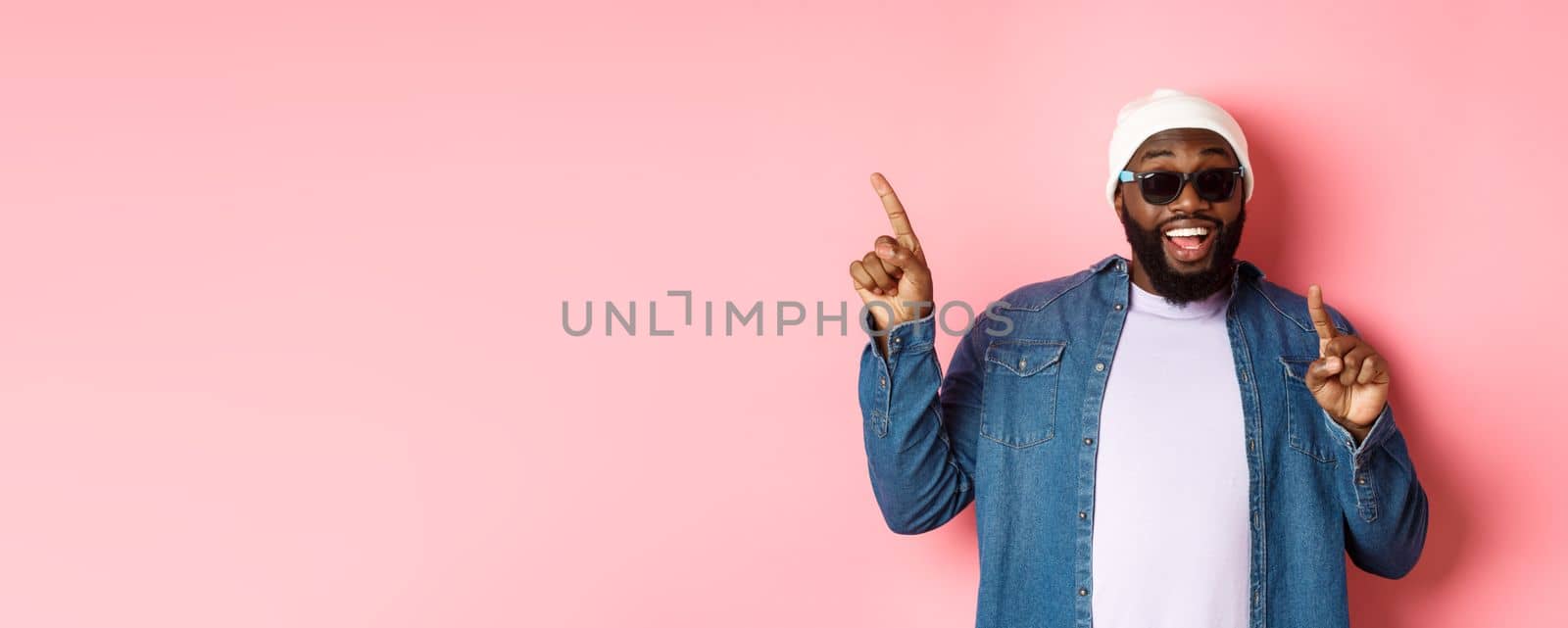 Happy bearded african-american man showing announcement, pointing fingers upper left corner and smiling, standing in beanie and sunglasses over pink background.