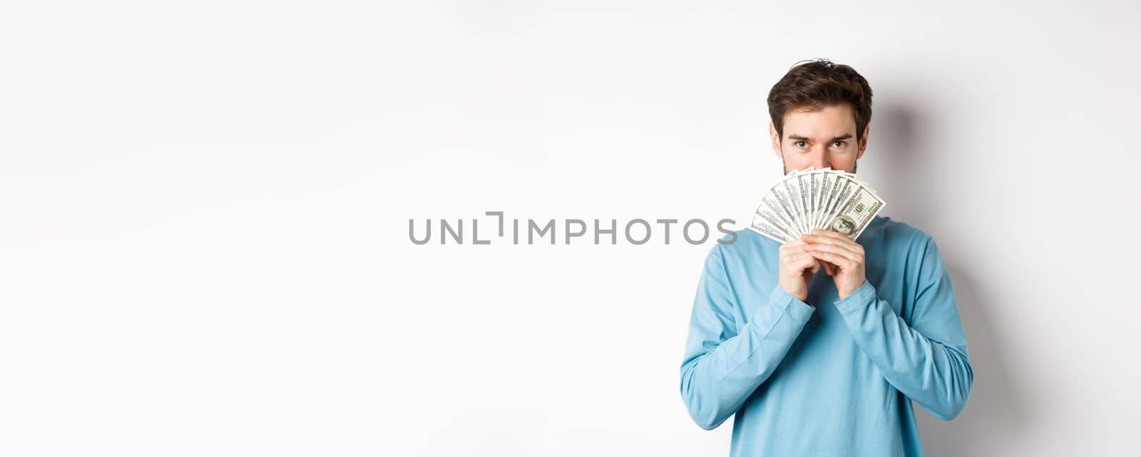 Finance concept. Smiling young man covering face behind money, showing dollars and looking happy, standing over white background by Benzoix