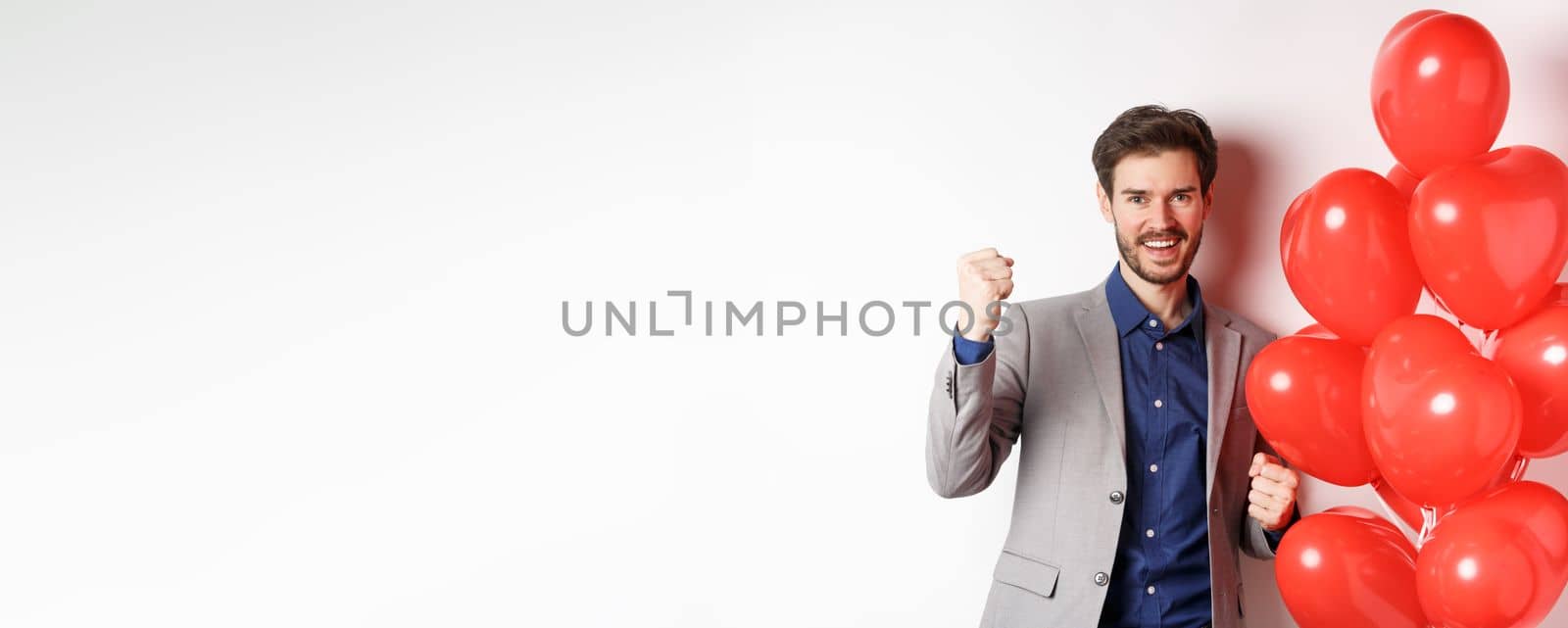 Lovers day. Cheerful young man in suit, feeling confident about Valentines date, saying yes and smiling, standing near hearts balloon, white background.