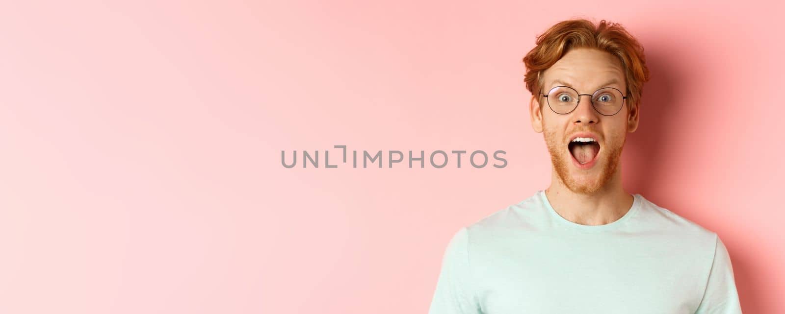 Close up of surprised redhead man raising eyebrows and scream with joy, checking out cool promo, looking amazed at camera, standing over pink background.