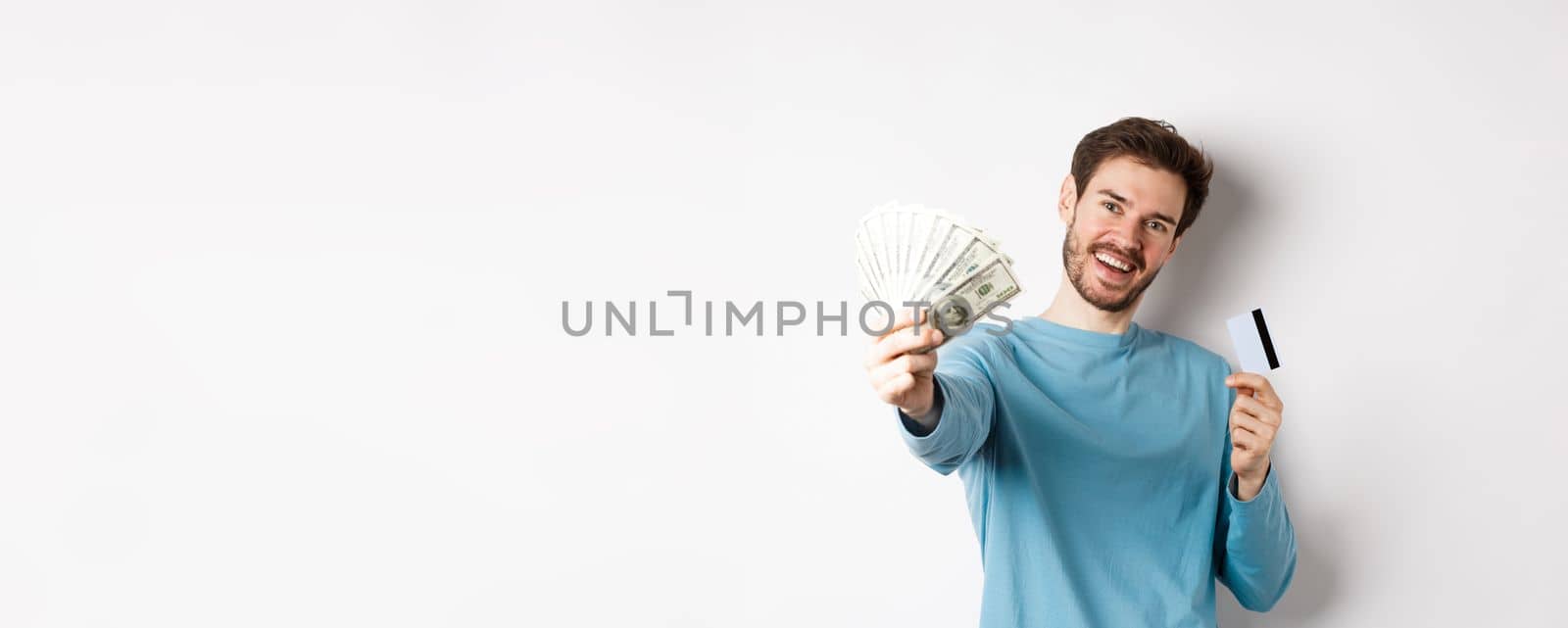 Handsome young man in blue shirt giving you money, holding plastic credit card, standing over white background by Benzoix