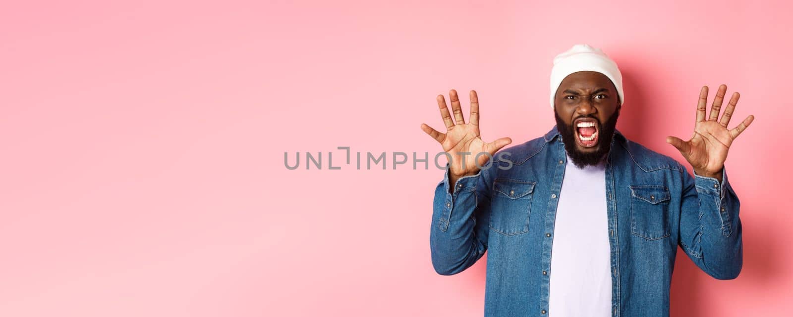 Angry african-american guy in beanie, scare you, roaring and screaming, showing hands, standing ove rpink background.