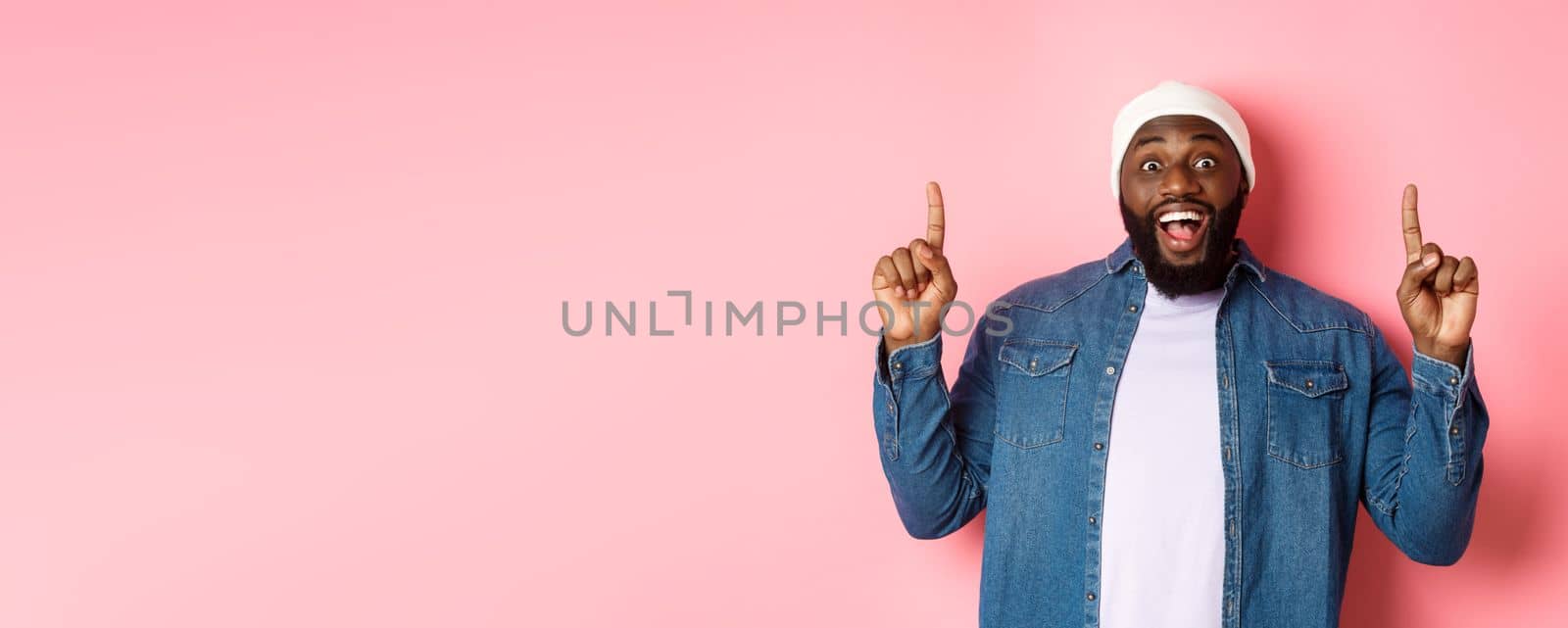 Cheerful Black man showing fantastic promo offer, pointing fingers up and smiling amused, standing over pink background by Benzoix
