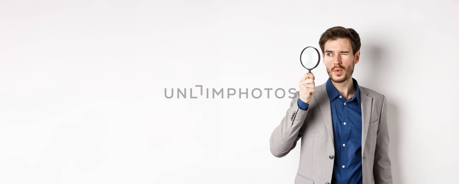 Handsome male model in suit looking through magnifying glass with interest, seeing something aside, white background.