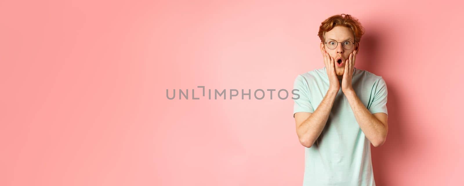 Shocked redhead man gossiping, staring impressed at camera and touching face, standing in glasses against pink background by Benzoix