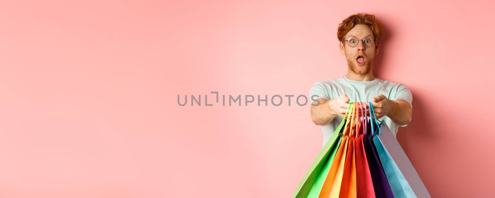 Surprised redhead man stretch out hands with shopping bags, give you gifts, standing over pink background.
