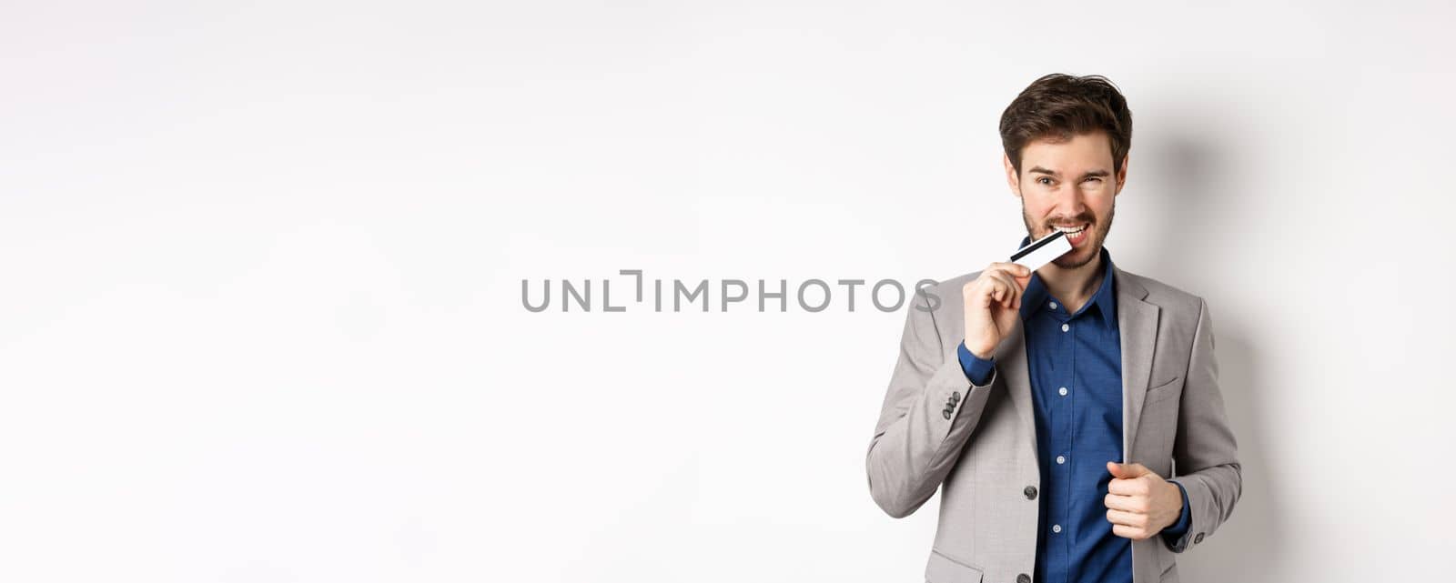 Successful young businessman in suit biting his credit card and looking sassy, standing on white background.
