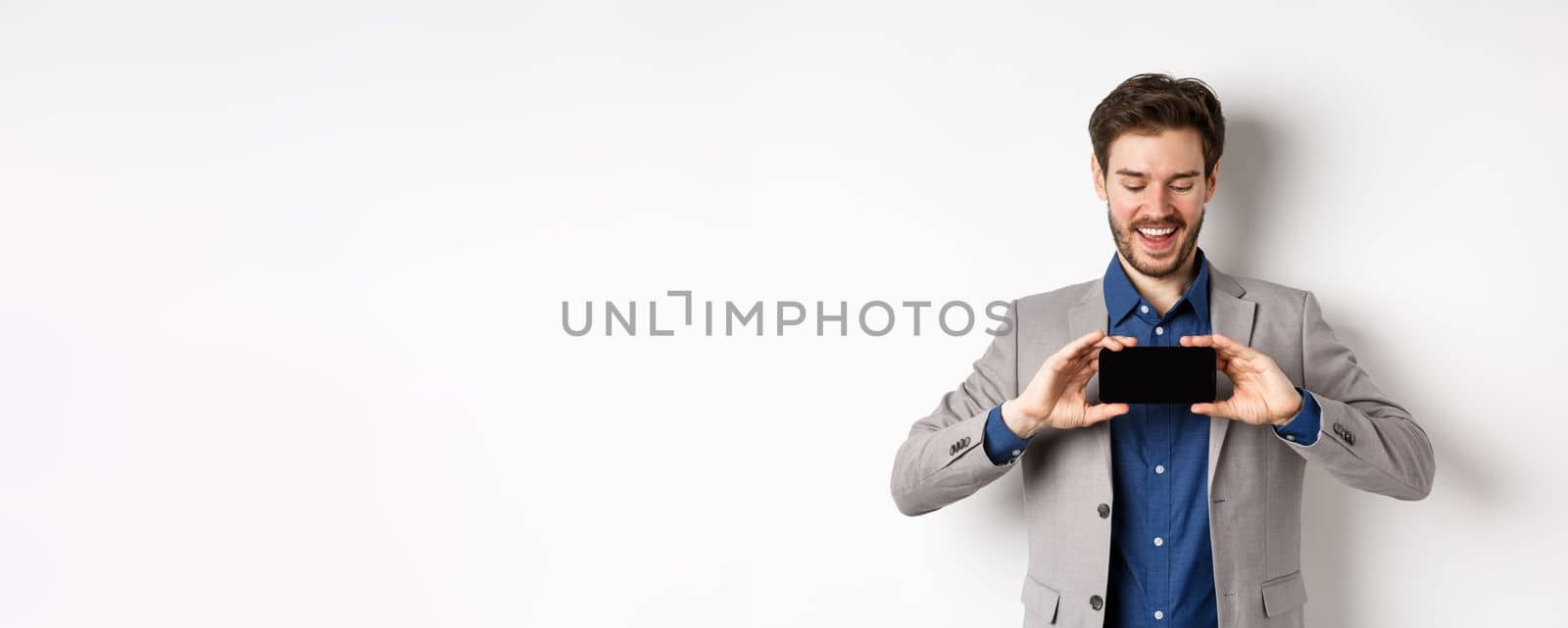E-commerce and online shopping concept. Happy young man in suit laughing, showing empty smartphone screen horizontally, wite background by Benzoix