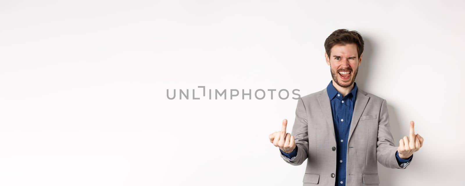Rude ignorant guy in business suit showing middle fingers and tongue, smiling while mocking people, fuck you gesture, standing on white background by Benzoix