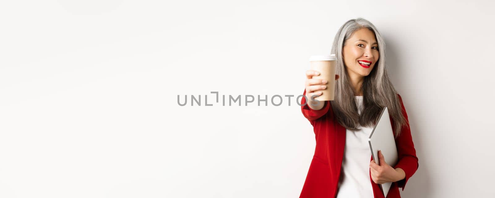 Business. Cheerful asian woman manager giving you cup of coffee and smiling, standing with laptop in hand, white background by Benzoix