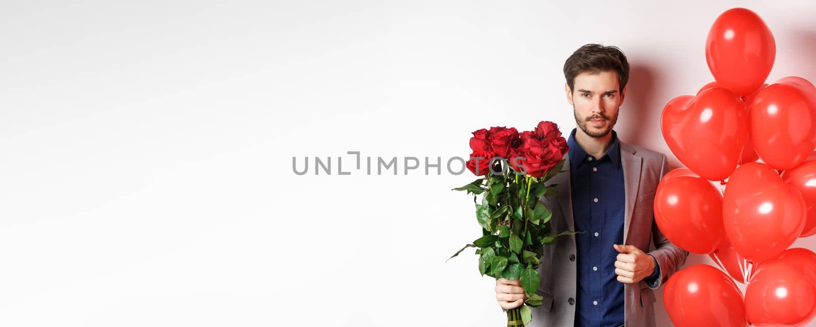 Valentines day. Handsome and confident man going on date in suit, holding bouquet of red roses and heart balloons, standing with gifts for lover on white background.