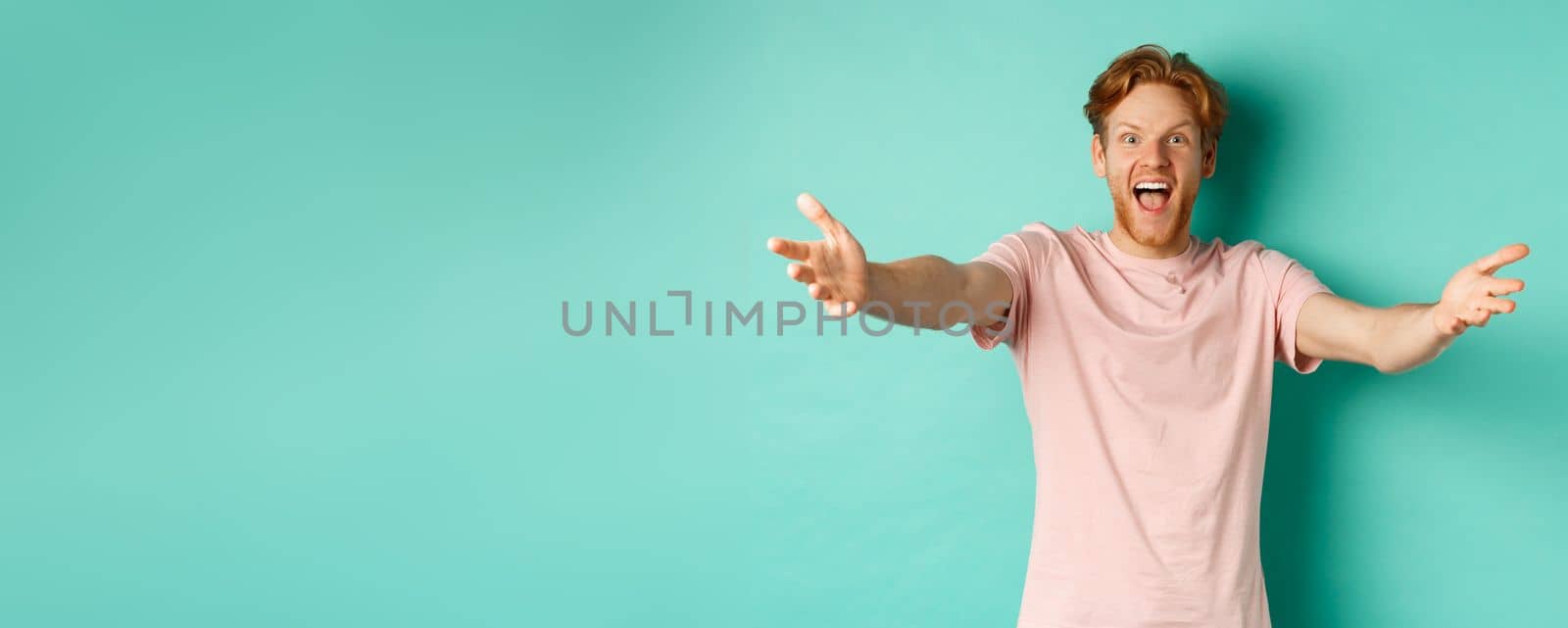 Friendly and happy young man with ginger hair, stretch out hands in warm welcome, reaching for hug and smiling joyfully, standing in t-shirt over mint background.