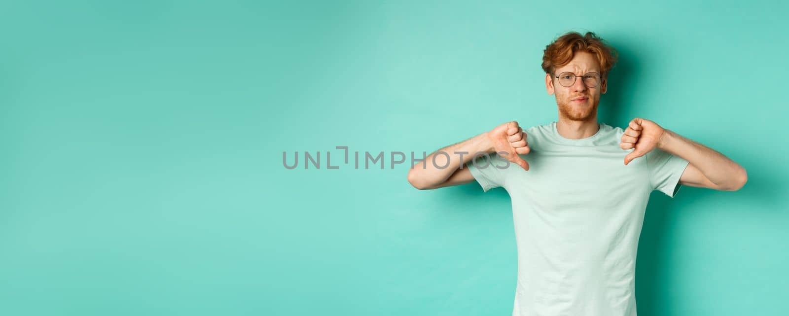 Disappointed caucasian guy with red messy hair and glasses showing thumbs down, dislike something, frowning upset and displeased, standing over mint background.