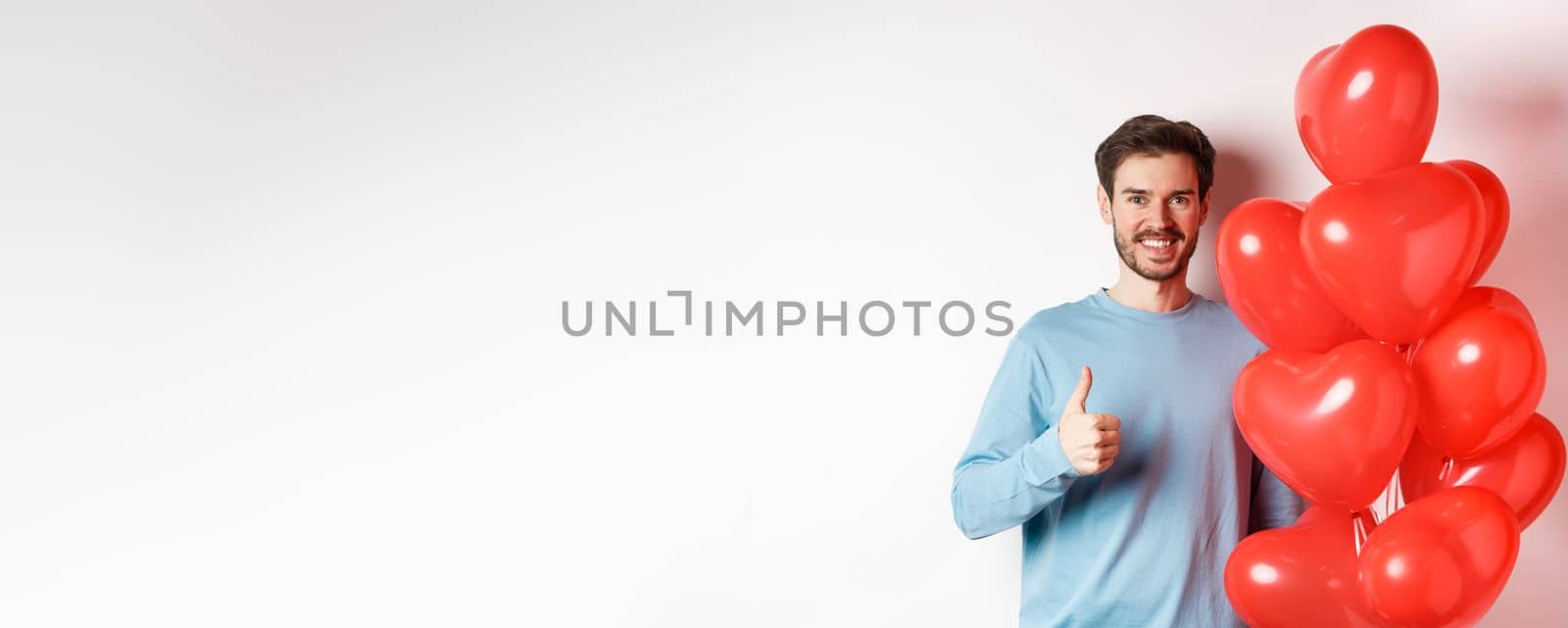 Smiling caucasian man standing with heart balloon, prepare surprise for lover on Valentines day, showing thumbs up and looking at camera, white background.