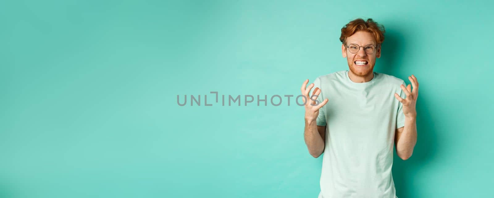 Portrait of distressed and angry redhead guy losing temper, shouting and shaking hands outraged, staring with furious face at camera, standing over mint background.