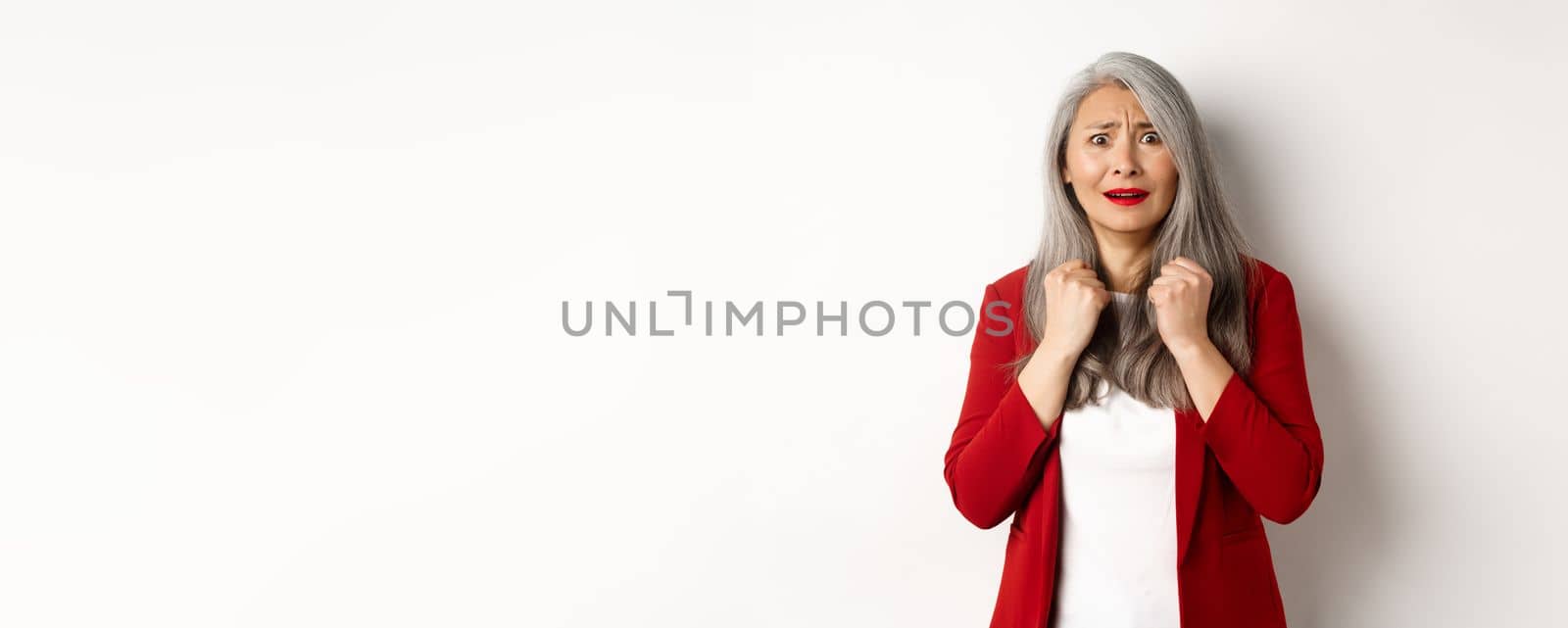 Business people. Scared mature asian woman looking terrified, trembling from fear, standing in red blazer over white background.