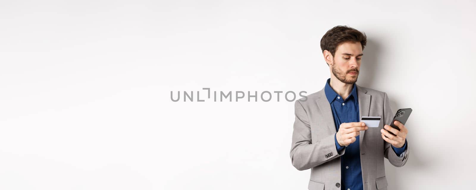 Online shopping. Serious man paying with credit card on smartphone, sending money, standing in suit on white background by Benzoix