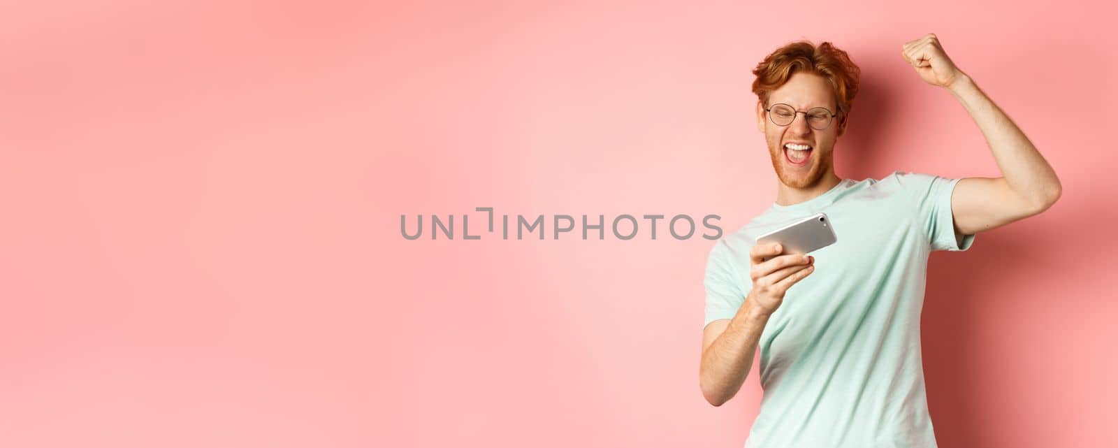 Happy redhead man winning in mobile video game, raising hand up and shouting yes with joy, celebrating victory, looking at smartphone, standing over pink background by Benzoix