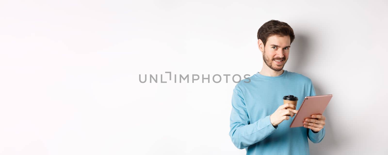 Handsome bearded man smiling at camera, drinking coffee and reading on digital tablet, standing over white background by Benzoix