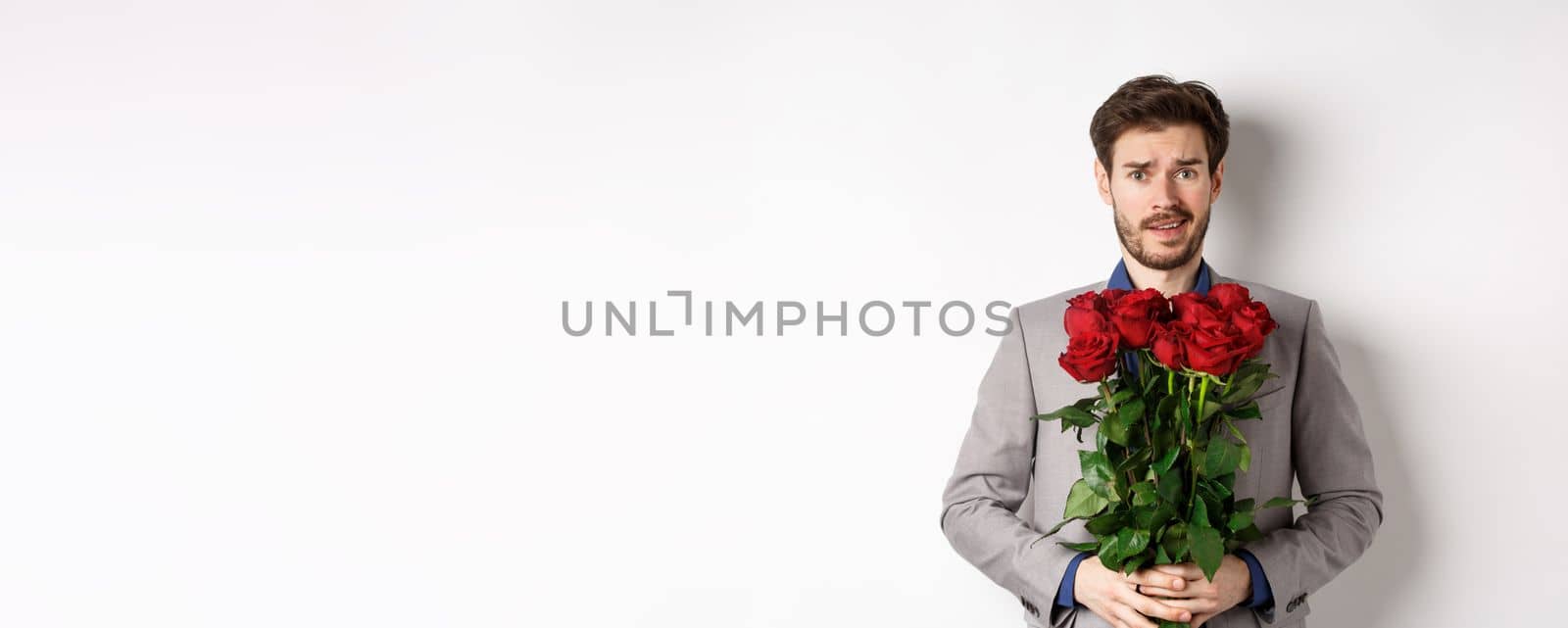 Worried boyfriend in suit, holding flowers roses and looking doubtful at camera, standing with bouquet on valentines day against white background.